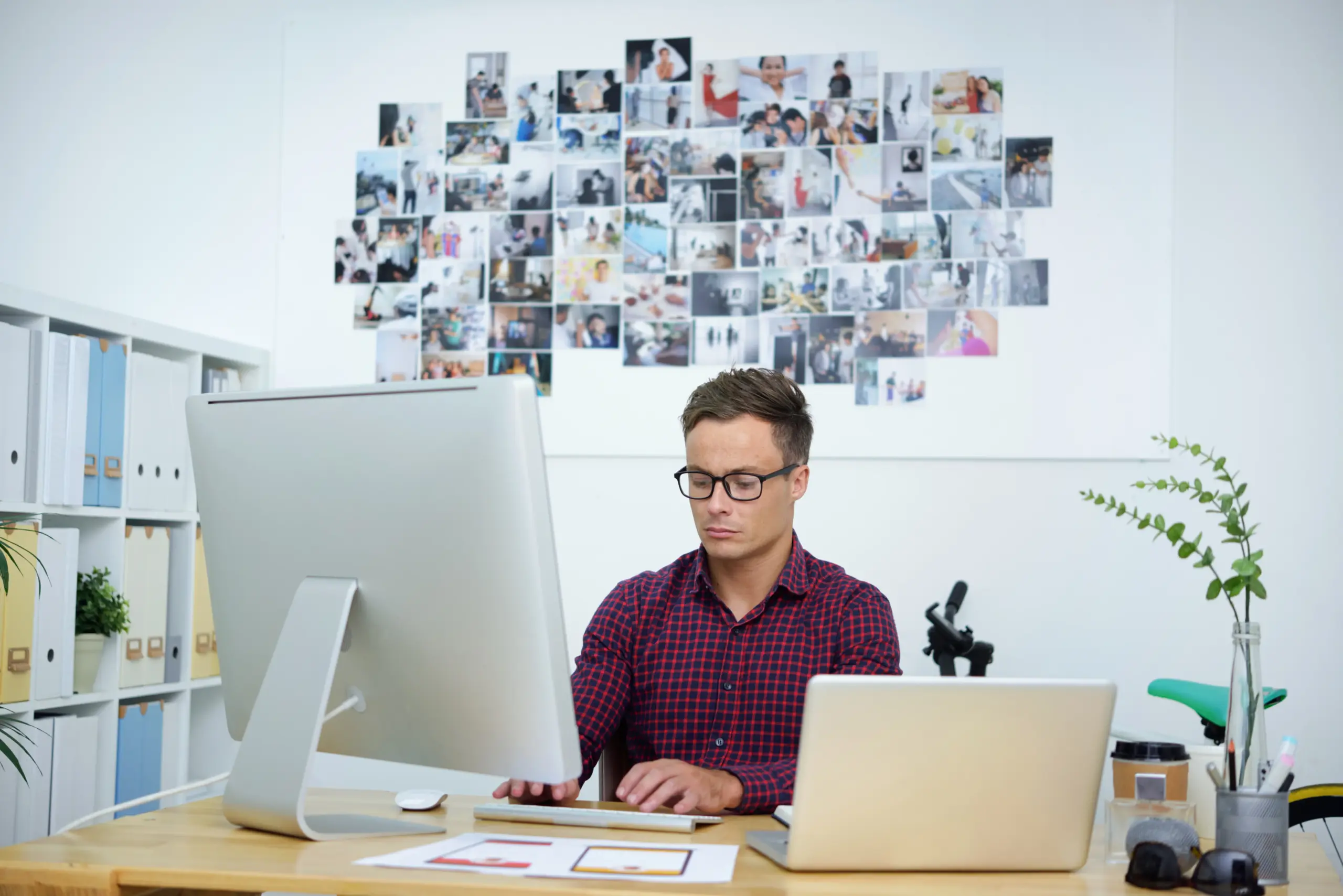 Man sitting at desk with two computers and pictures on the wall behind him.